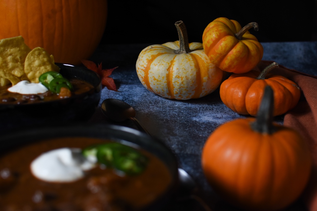 Colorful pumpkins in front of a dark background with pumpkin chili blurred in the foreground. 